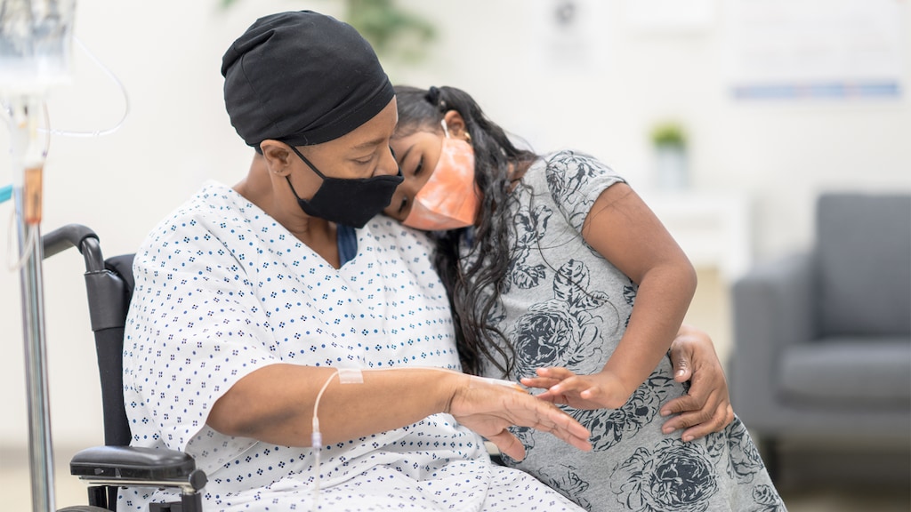 A mature woman in a wheelchair wearing a protective mask and headwrap shows her young daughter her IV
